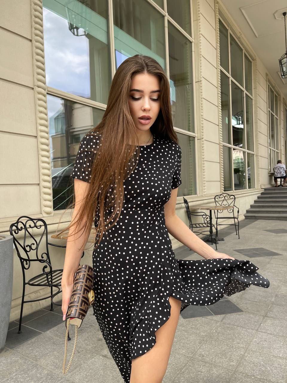 Woman in a black polka dot dress standing on a sidewalk with a building in the background