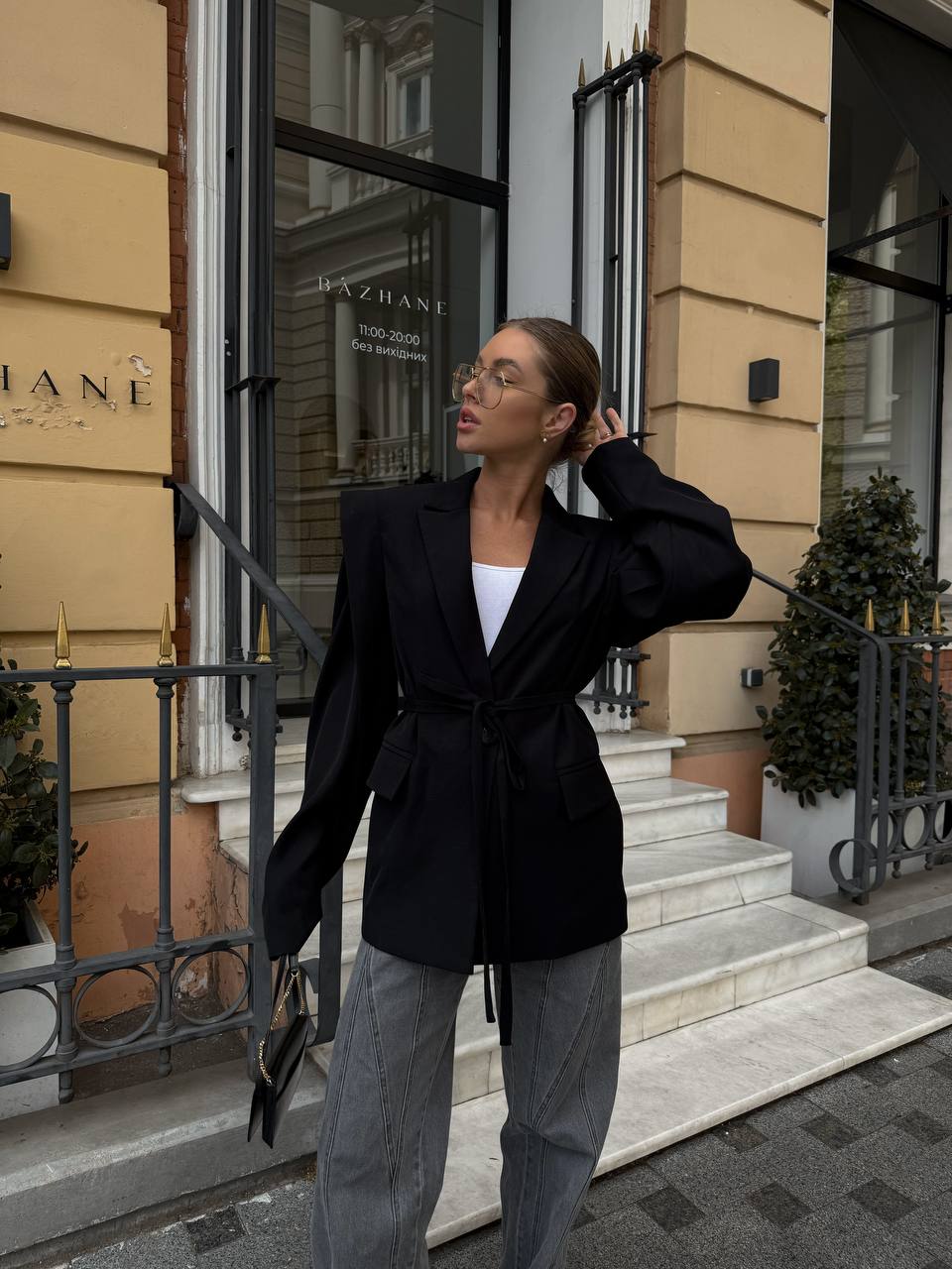 Woman in a black blazer and gray pants standing in front of a building entrance.