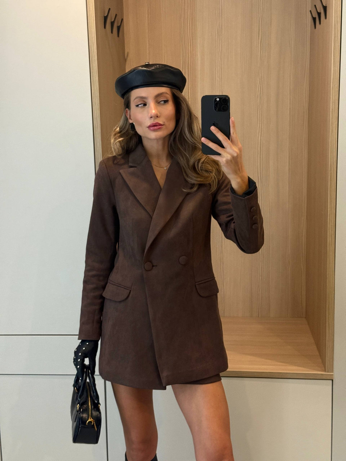 A woman takes a mirror selfie wearing a dark brown double-breasted blazer dress, a black beret, black gloves, and holding a black handbag with gold hardware.