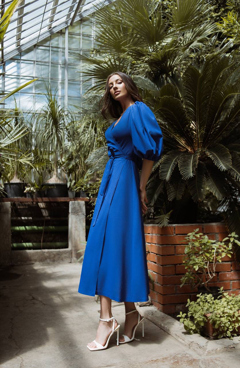 Woman in a blue dress standing among plants in a greenhouse.