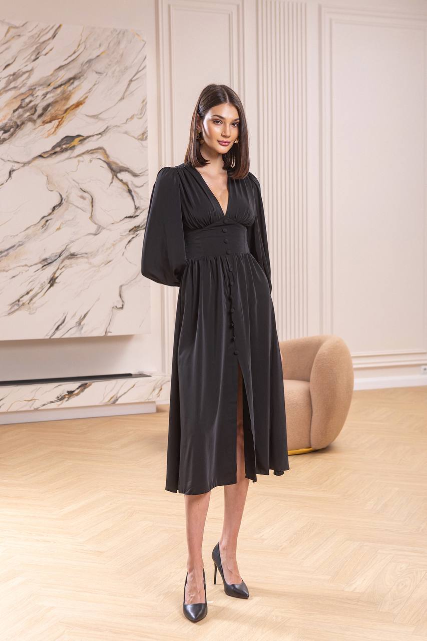 Woman in a black dress standing in a stylish room with marble wall and beige armchair.