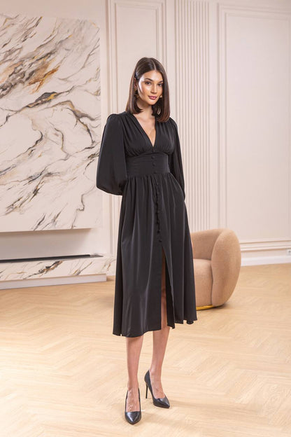 Woman in a black dress standing in a stylish room with marble wall and beige armchair.