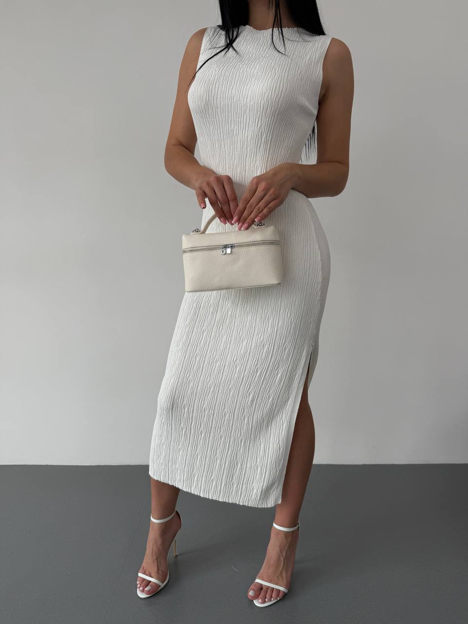 Woman wearing a white sleeveless dress holding a matching handbag against a gray background