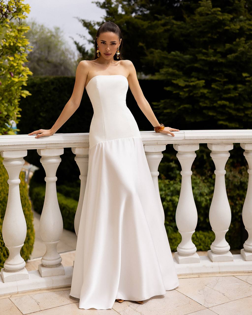 Woman in a white strapless gown standing on a balcony with greenery in the background