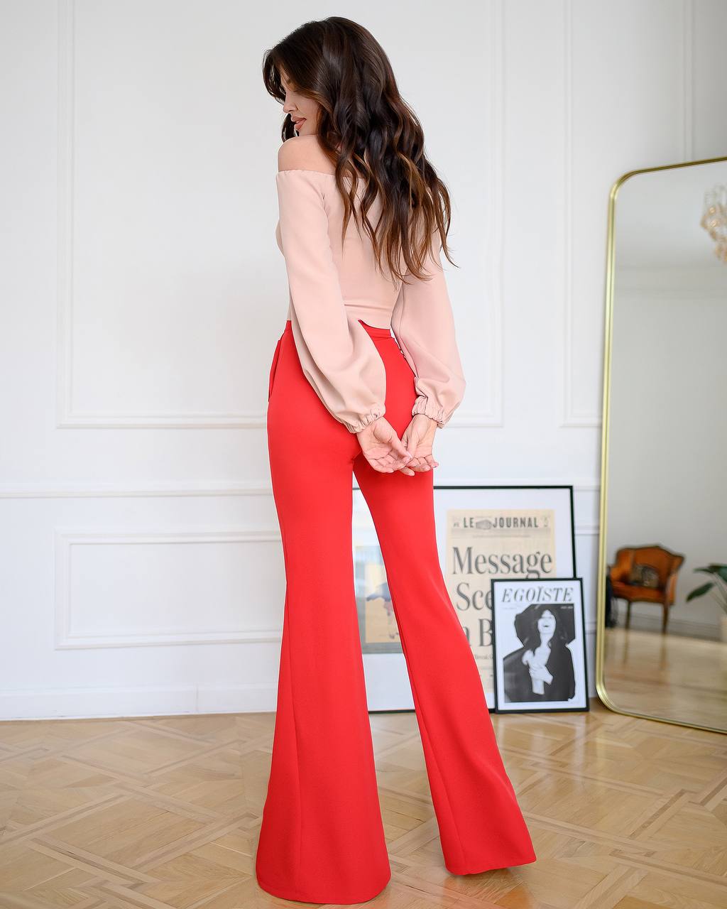 Woman wearing a light pink top and red flared pants standing in a room with a mirror and decor.