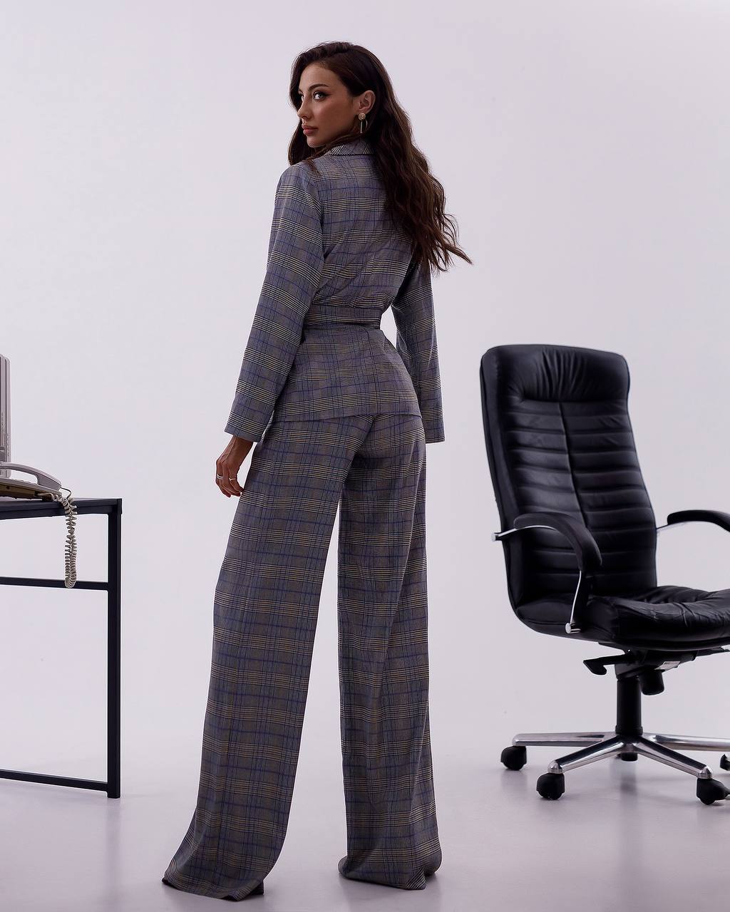 Woman in a plaid suit standing in an office setting with a black chair and desk.