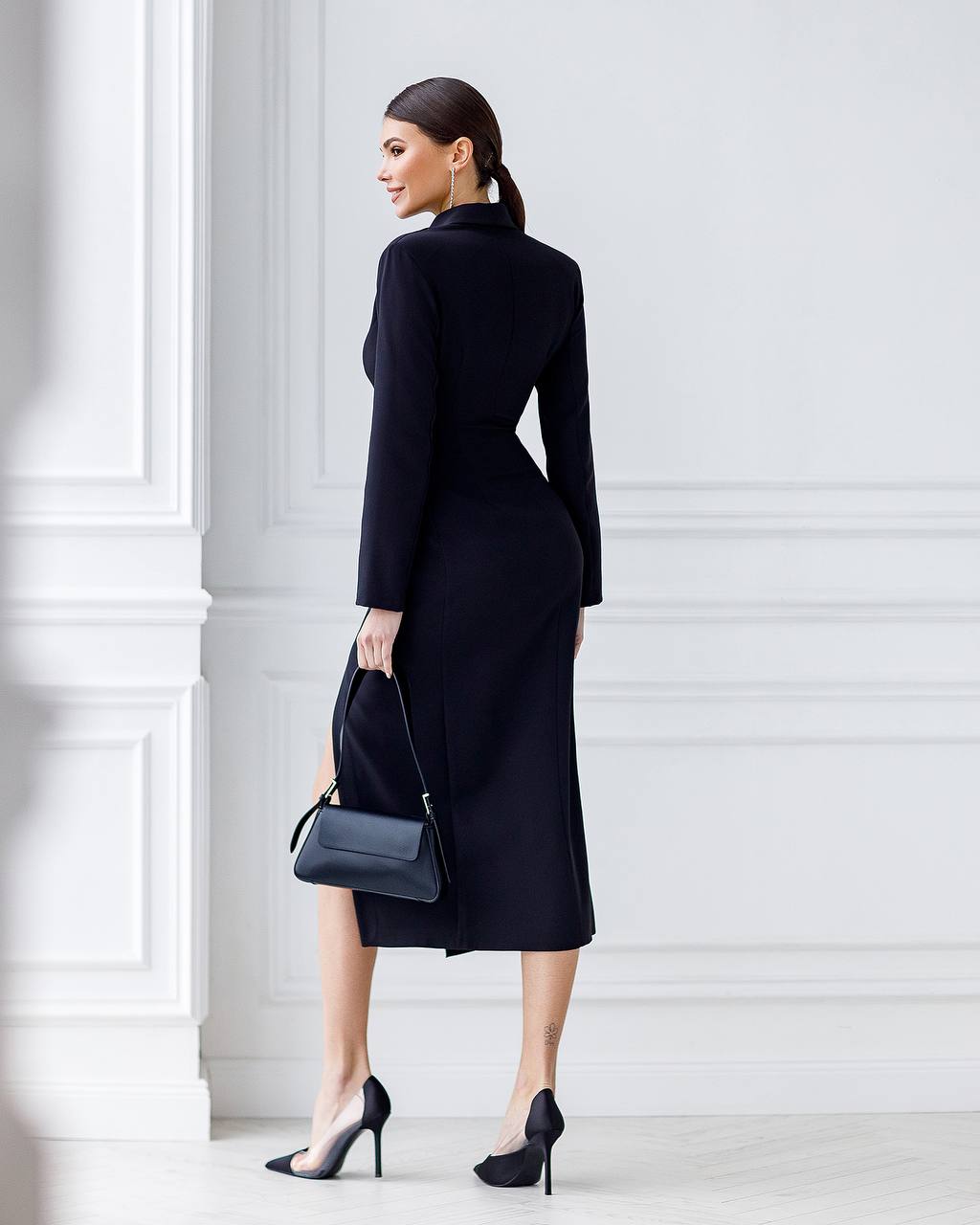 Woman wearing a black coat and holding a black handbag against a white wall.
