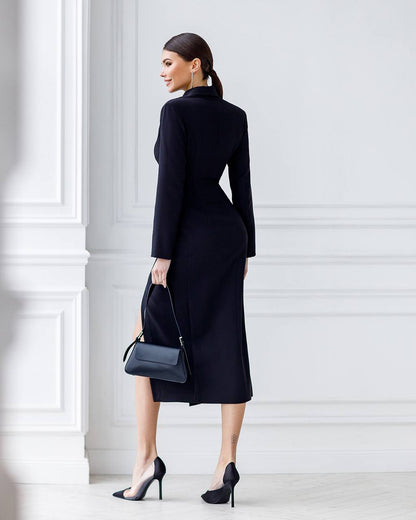 Woman wearing a black coat and holding a black handbag against a white wall.