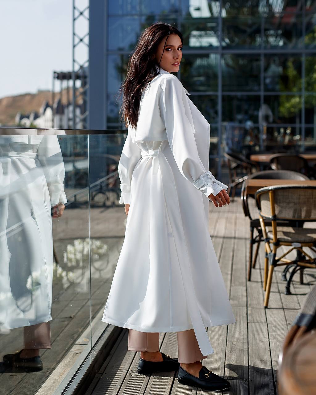 Woman in a white dress standing on a wooden deck with glass walls.