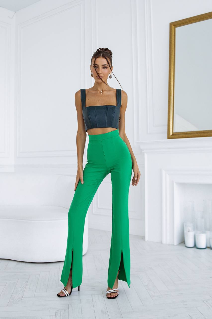 Woman wearing a black top and green pants in a room with white walls and a mirror.