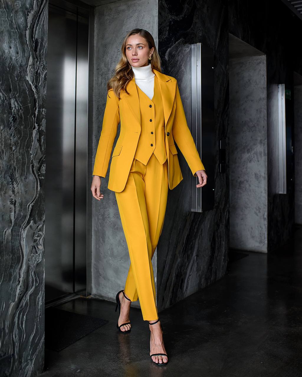 Woman in a yellow suit standing in a modern, darkly lit interior setting.