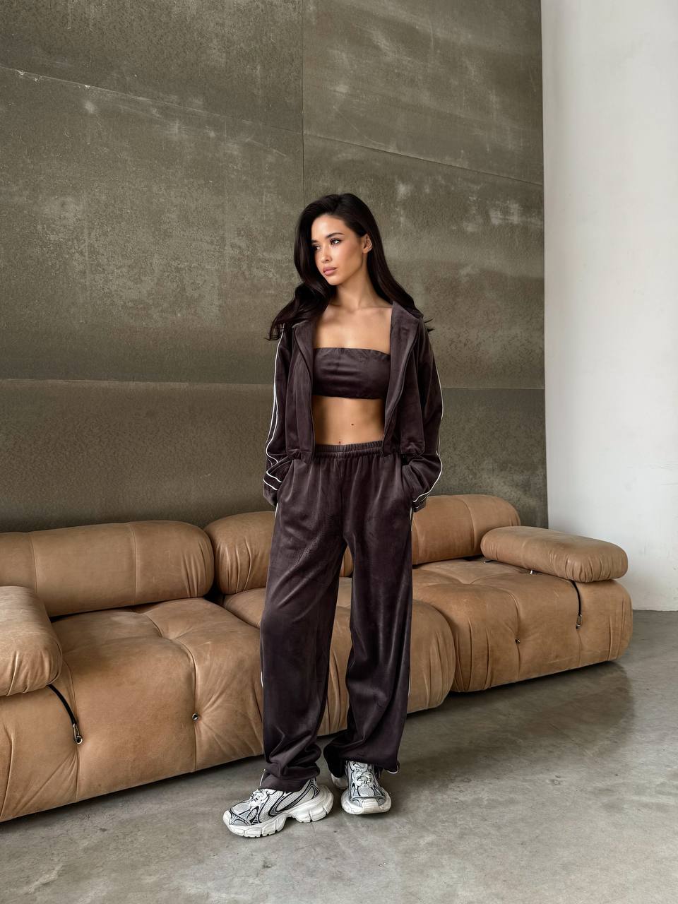 Woman in a brown tracksuit standing in front of a brown sofa against a gray wall.