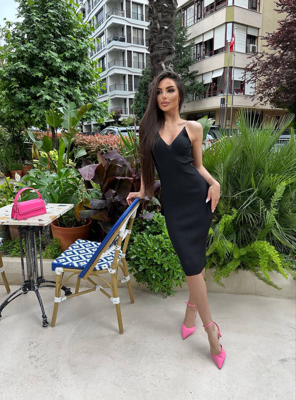 Woman in a black dress with pink shoes standing outdoors near greenery and a building.