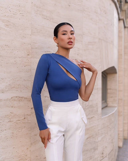Woman wearing a blue top with a cutout design and white pants against a beige wall.