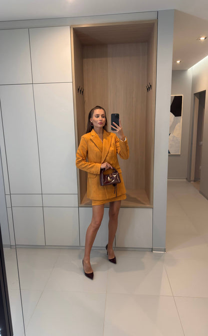 A woman takes a mirror selfie wearing a mustard yellow tailored blazer and matching mini skirt set, holding a dark brown handbag and wearing dark pointed-toe heels in a modern interior setting.