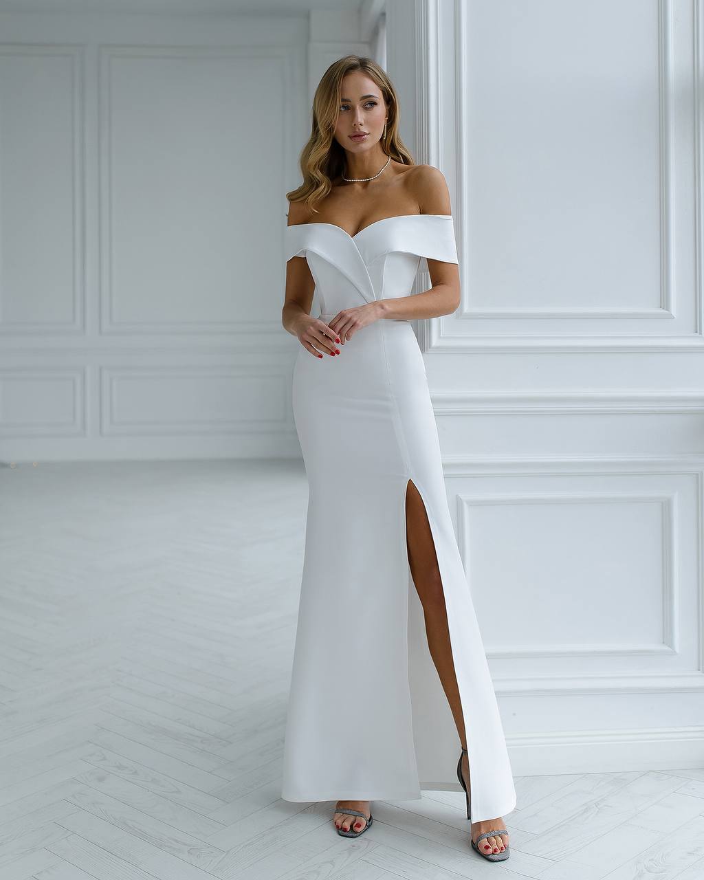 Woman wearing a white off-shoulder gown with a slit in a minimalistic room.