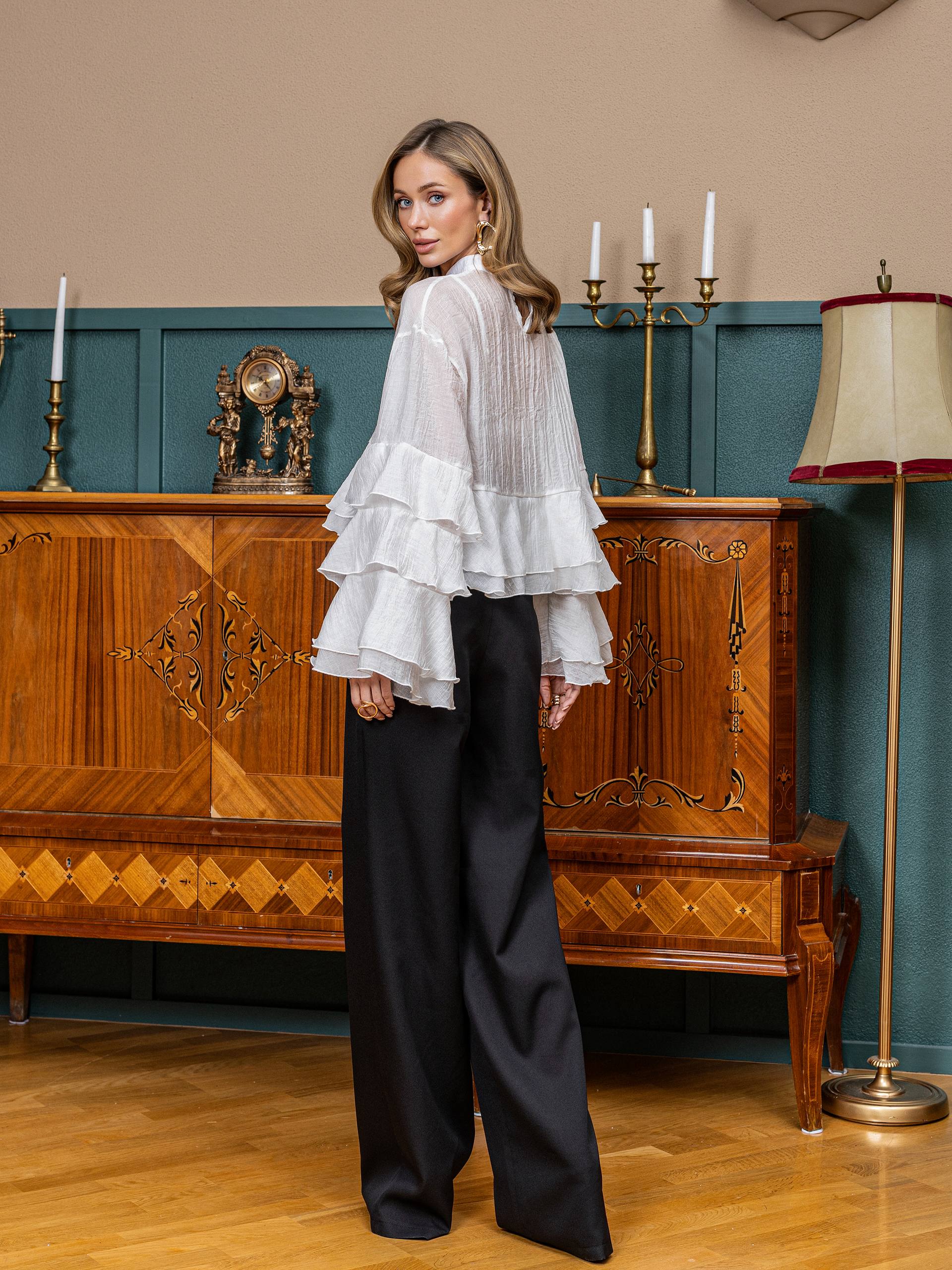 Woman in a white ruffled blouse and black pants standing in a room with wooden furniture and decorative elements.