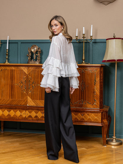 Woman in a white ruffled blouse and black pants standing in a room with wooden furniture and decorative elements.