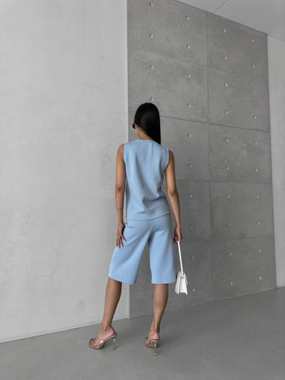 Woman in a light blue sleeveless top and shorts standing against a gray concrete wall.