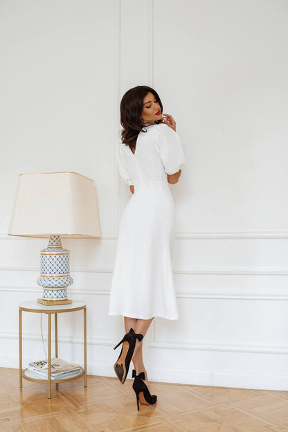 Woman in a white dress standing in a room with a lamp and magazine on a table.
