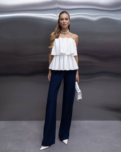 A woman models a white off-the-shoulder ruffled peplum top paired with navy blue wide-leg trousers and white pointed shoes, standing against a metallic gray background.