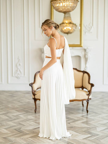 A woman wearing a white two-piece pleated dress with a crop top and long draped veil, standing in a bright white room with a chaise lounge and chandelier in the background.