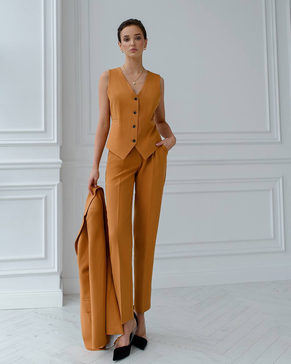 Woman wearing a mustard yellow suit standing against a white paneled wall.