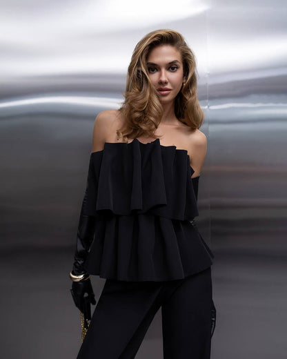 A woman modeling a black off-the-shoulder ruffled top with matching black pants and gloves, posed against a dark, abstract metallic background.
