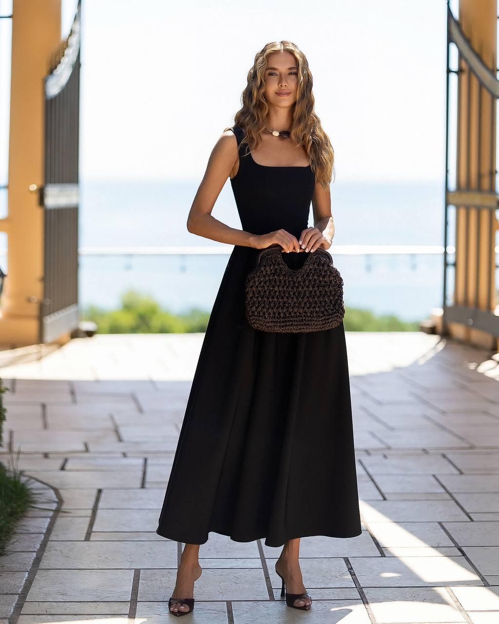 A woman stands on a tiled outdoor walkway holding a dark brown woven handbag. She is wearing a sleeveless black midi dress and black heels, with a bright, sunlit background showing a blurred coastal view.