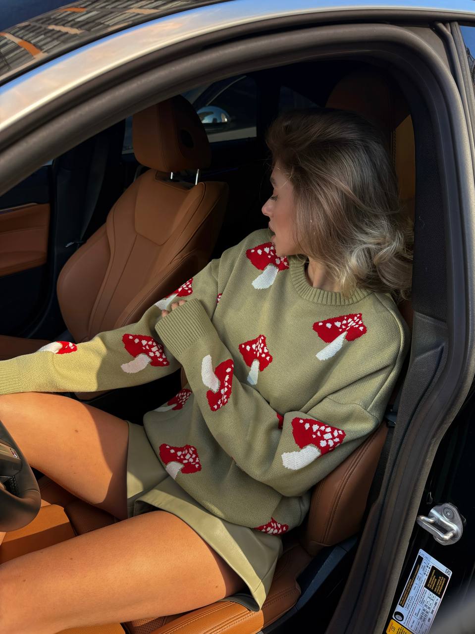 Woman sitting in a car wearing a green sweater with red and white mushroom patterns.