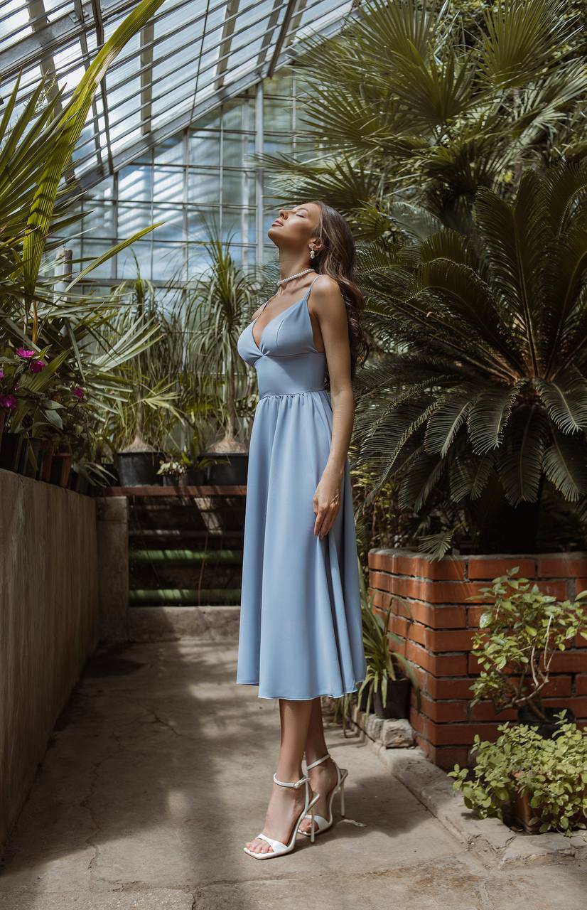 Woman in a light blue dress standing in a greenhouse filled with plants.
