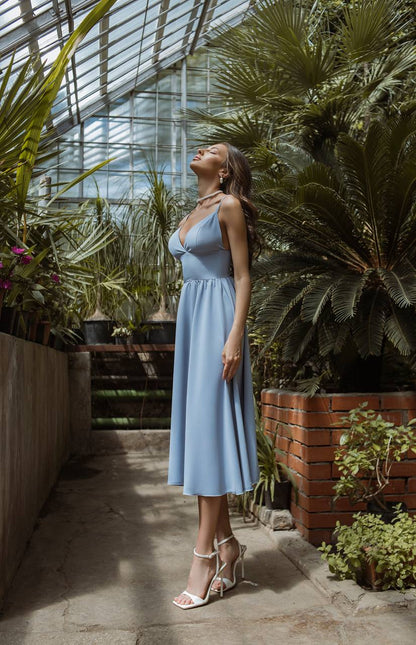 Woman in a light blue dress standing in a greenhouse filled with plants.