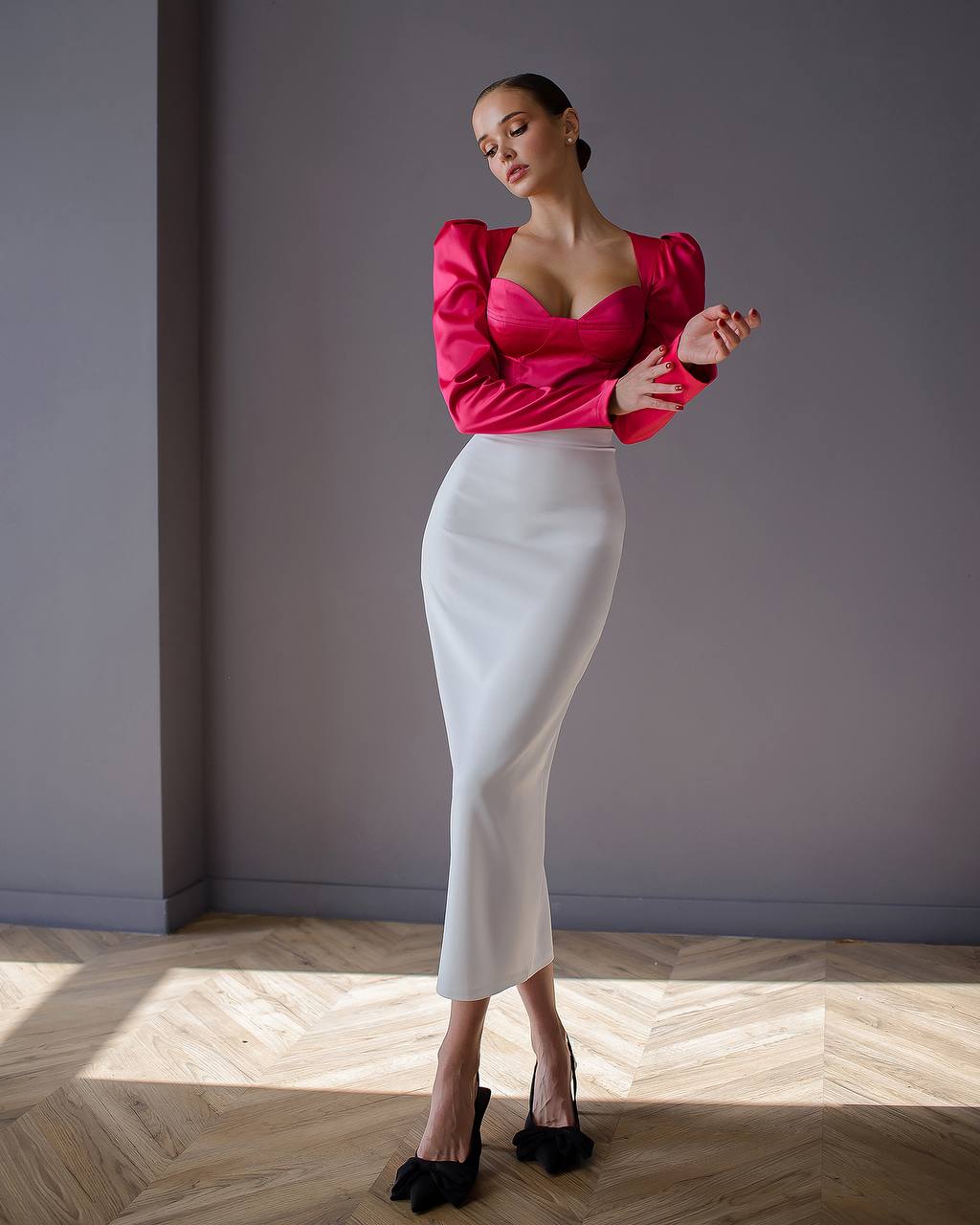 Woman wearing a pink top and white skirt standing against a gray wall.