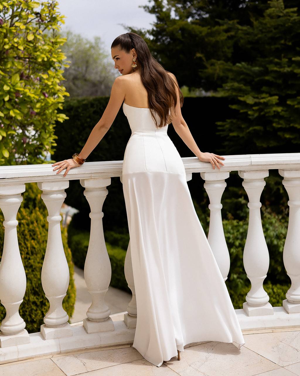 Woman in a white strapless gown standing on a balcony with greenery in the background