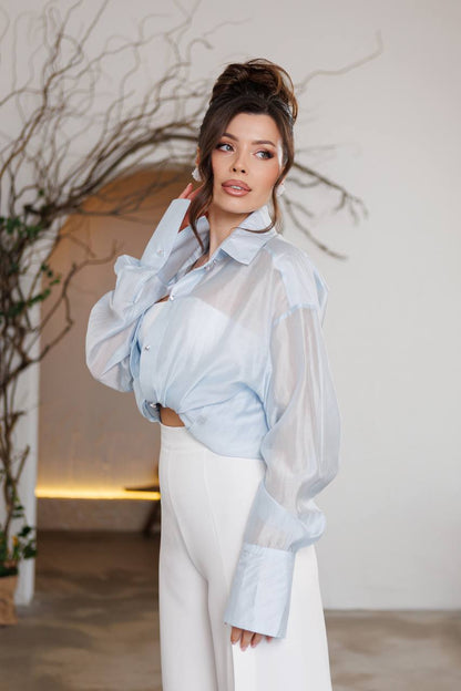 Woman wearing a light blue sheer blouse over a white outfit with a neutral background