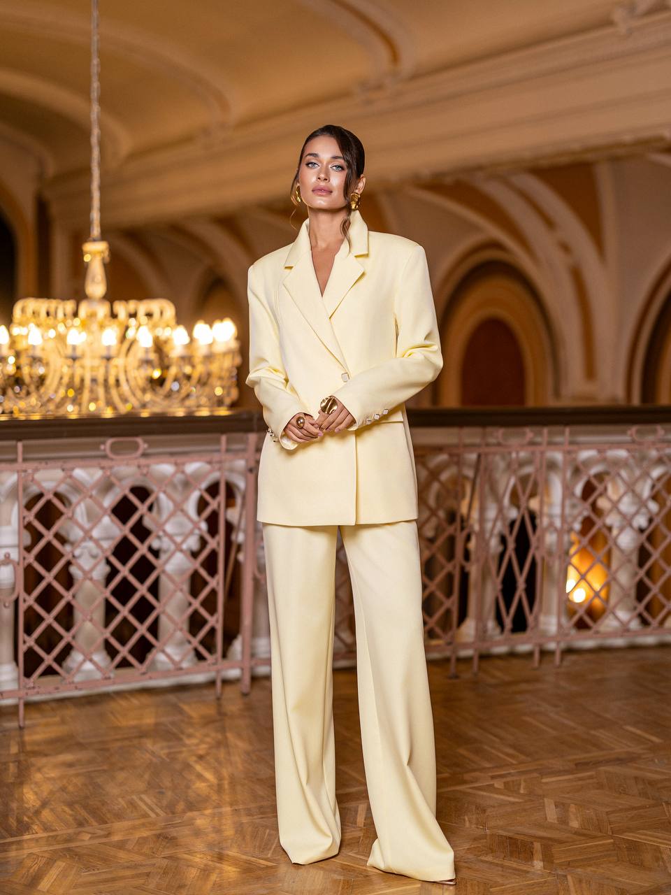 Woman in a light-colored suit standing in an elegant interior setting with chandeliers.
