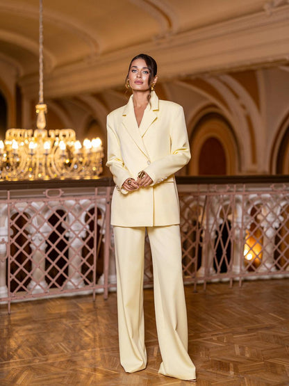 Woman in a light-colored suit standing in an elegant interior setting with chandeliers.