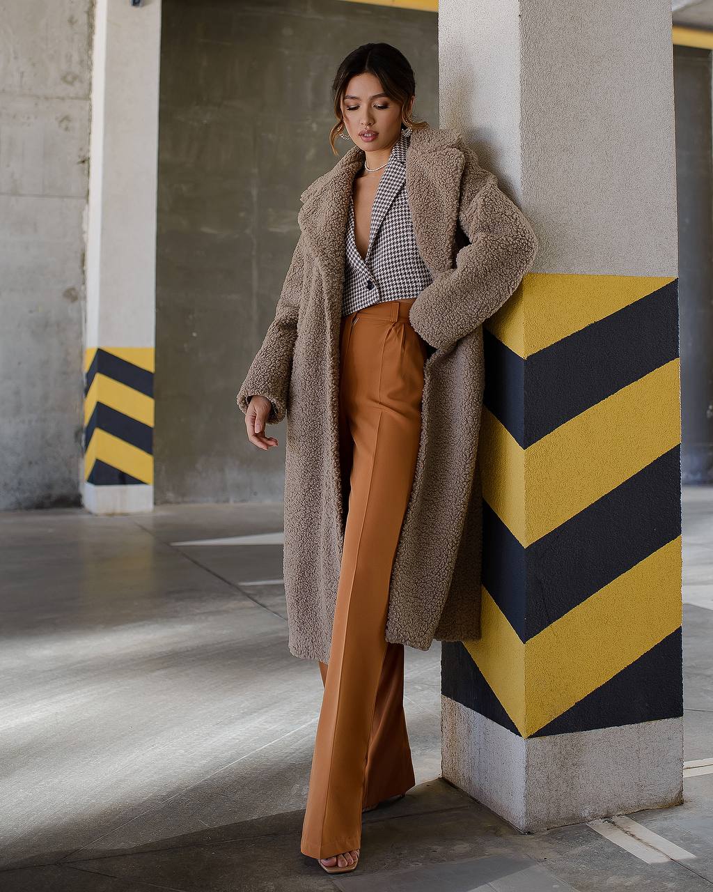 Woman in a long brown coat and mustard pants standing against a concrete wall with yellow and black stripes.
