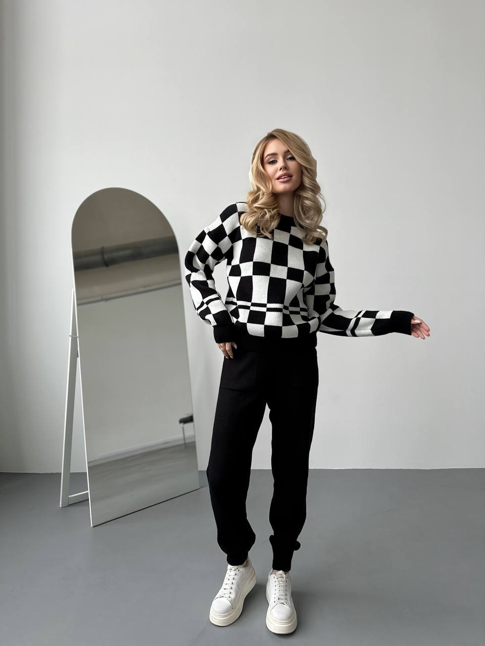 Woman wearing a black and white checkered sweater and black pants standing in front of a mirror.