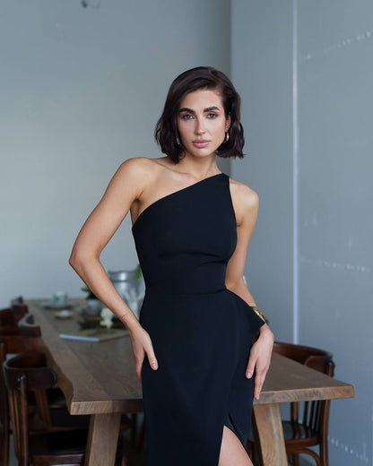 Woman in a black one-shoulder dress standing in a room with wooden furniture.