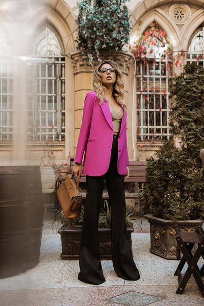 Woman in a pink blazer and black pants standing in an elegant outdoor setting with plants and architectural elements.