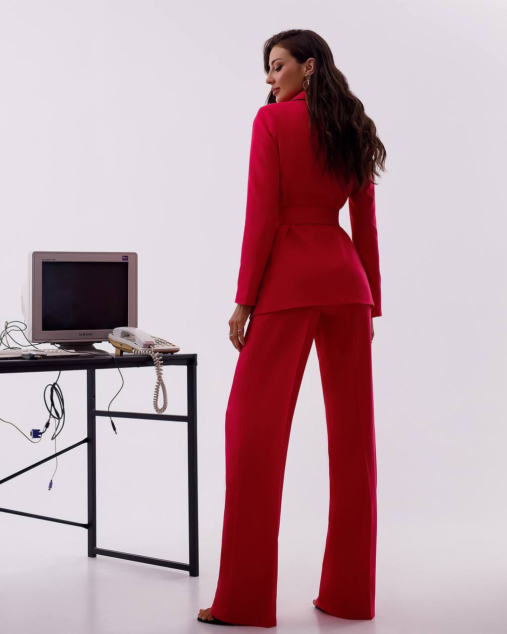 Woman in a red suit standing in front of a desk with a computer monitor and phone.