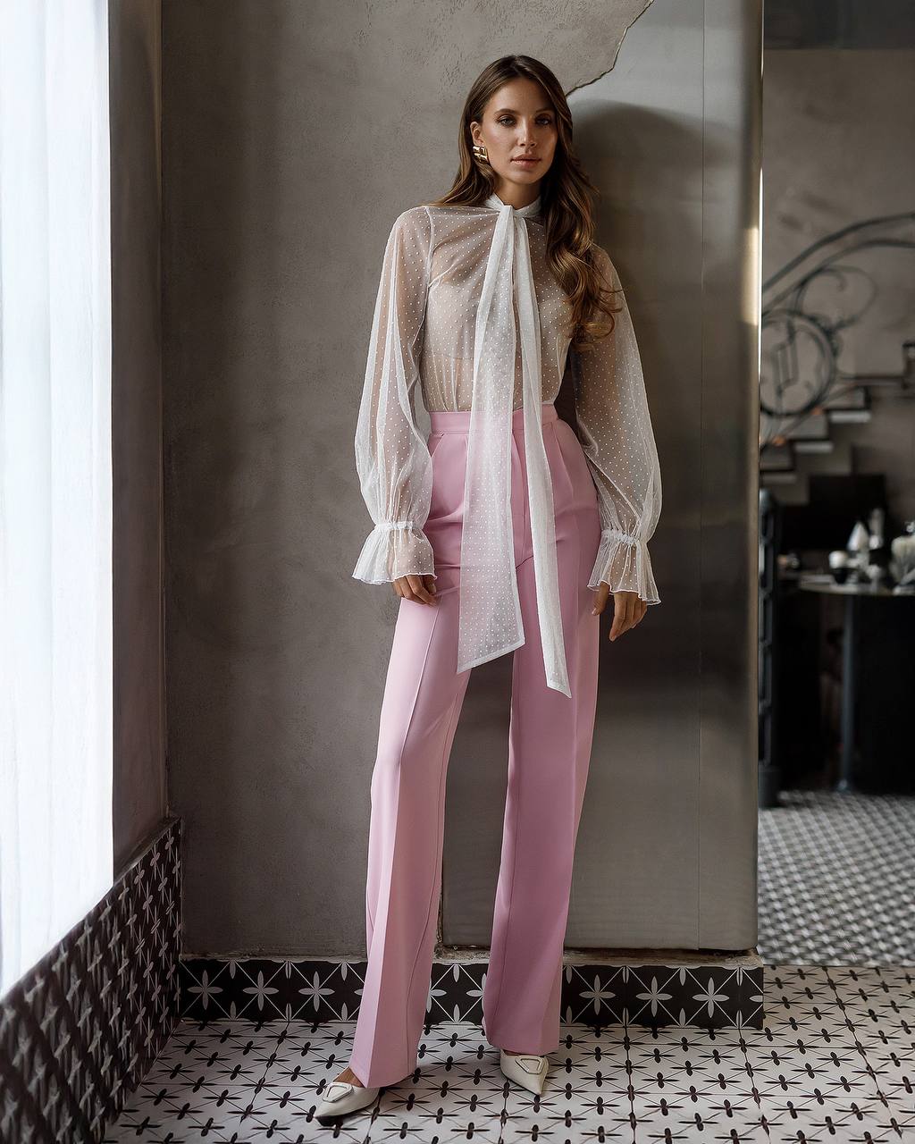 Woman wearing a light blouse and pink pants standing against a textured wall.