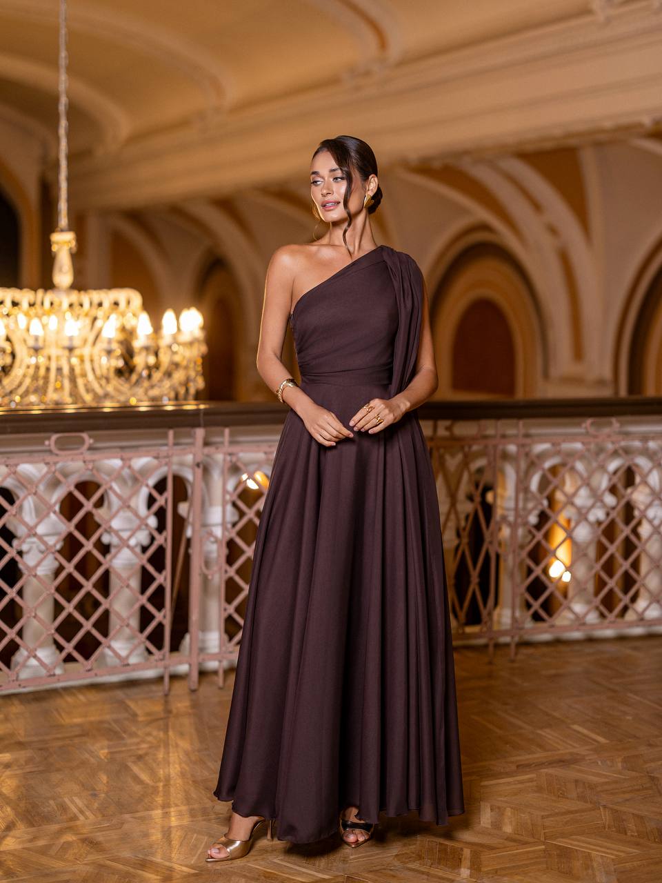 Woman in a dark purple one-shoulder gown standing in an elegant interior setting with chandeliers.