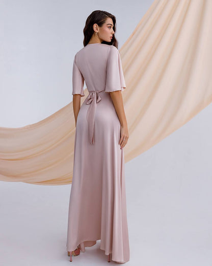 Woman wearing a long, light pink dress with a soft fabric draped behind her on a plain background