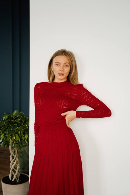 Woman wearing a red dress standing against a white wall with a plant to her left.