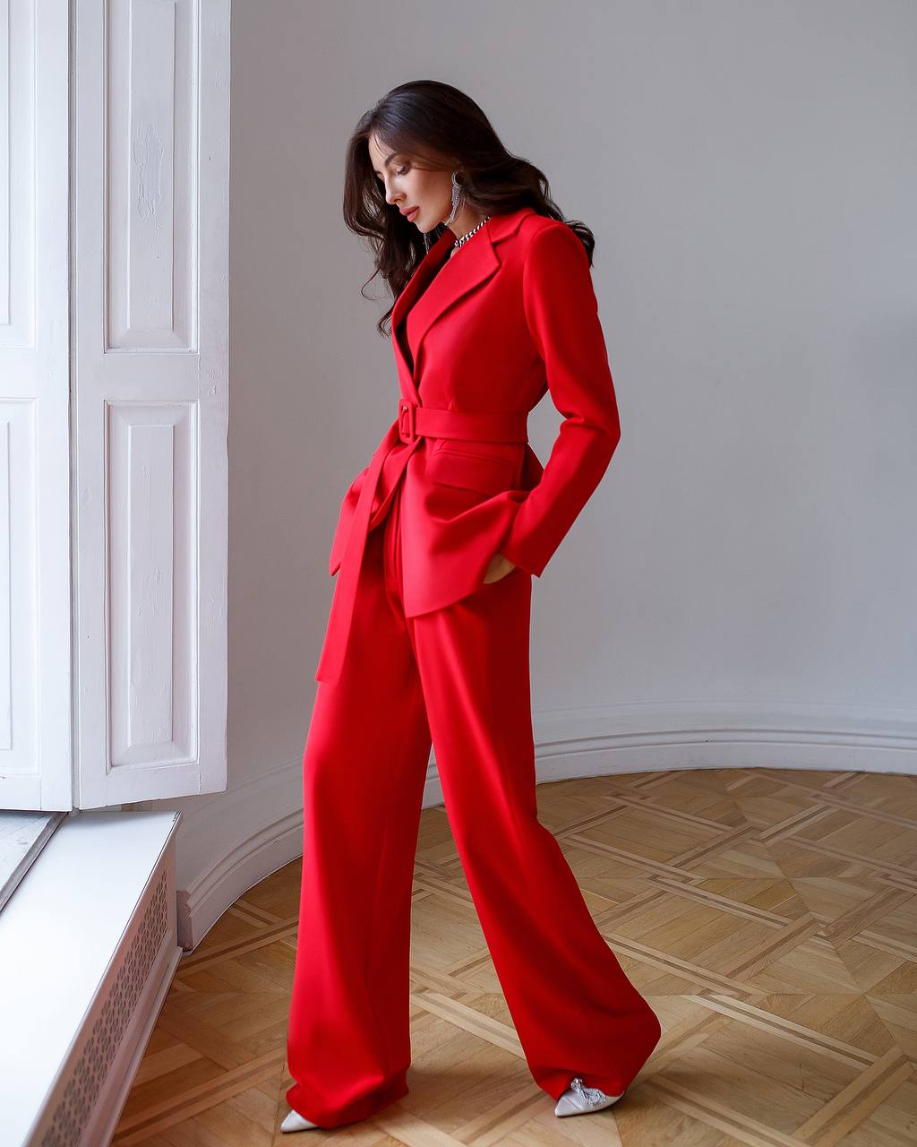 Woman wearing a red suit standing in a room with wooden flooring and white walls.