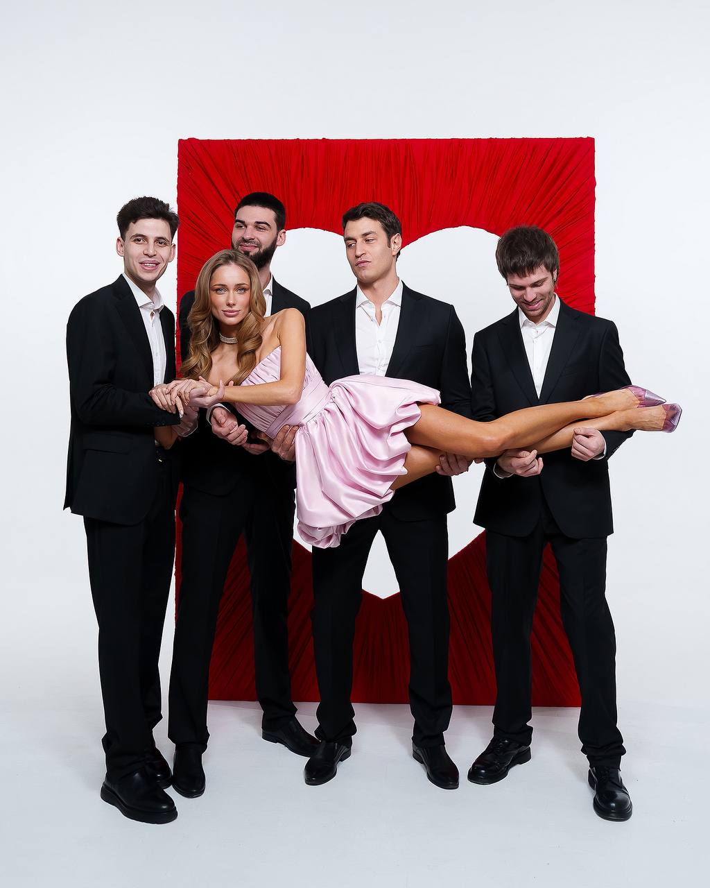 Four men in suits holding a woman in a pink dress against a red curtain background