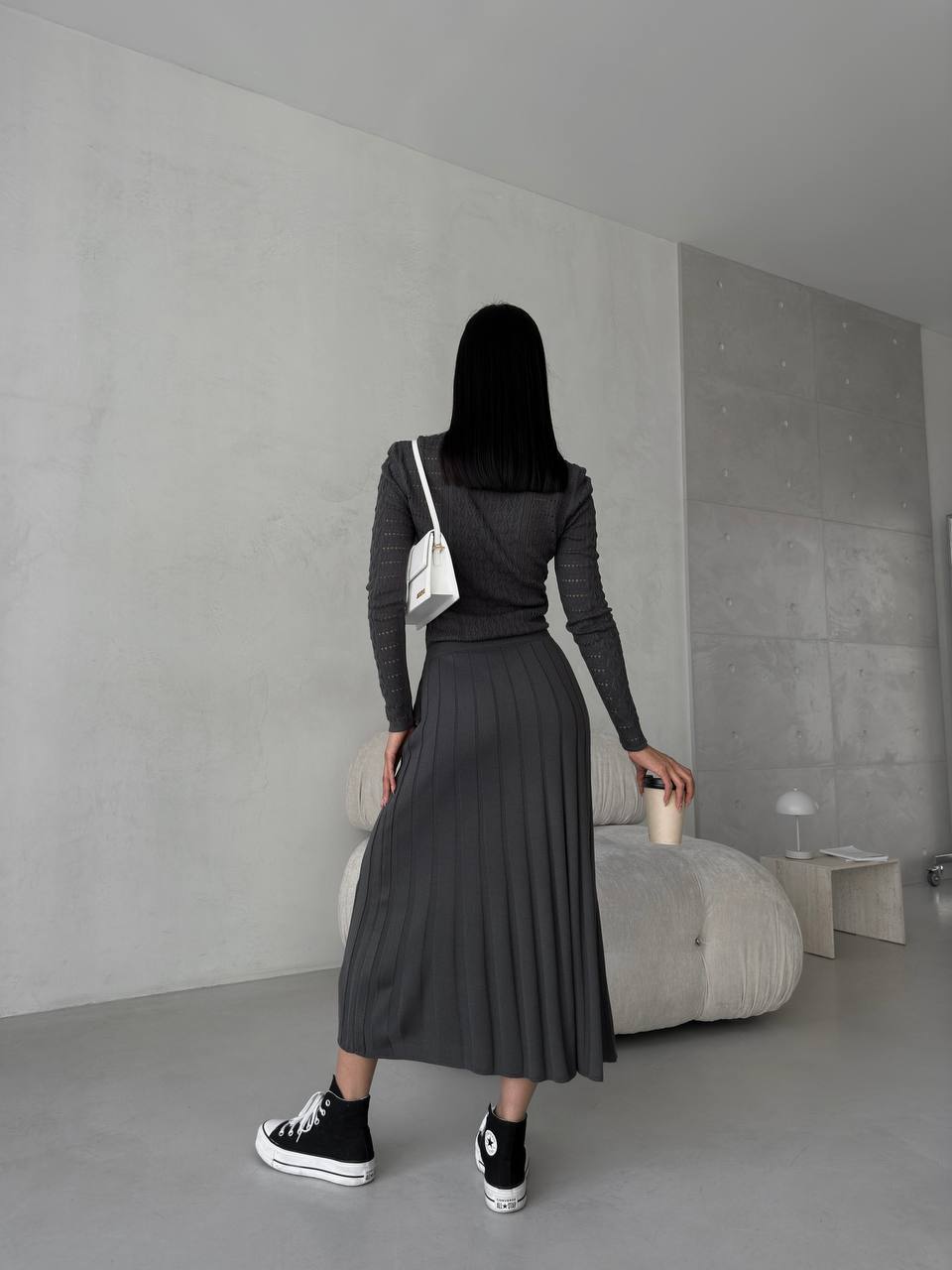 Person wearing a dark outfit with a white bag in a minimalistic room.