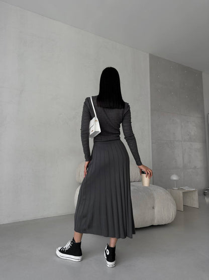 Person wearing a dark outfit with a white bag in a minimalistic room.